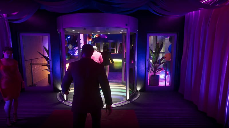 A man in a dark suit, seen from behind, approaches a lit revolving door at the Megamundo Building, with balloons and plants inside and purple curtains around it.