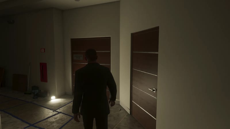 A man in a dark suit, seen from behind, walks along a dim corridor towards a wooden double door and a single door in the Megamundo Building.