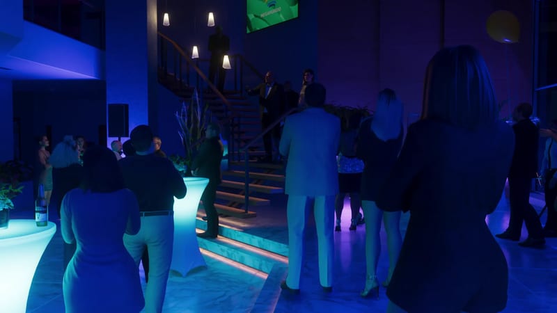 Guests gather at a party in a blue-lit atrium beneath a staircase, with a Megamundo logo on a green screen above, in the Megamundo Building.