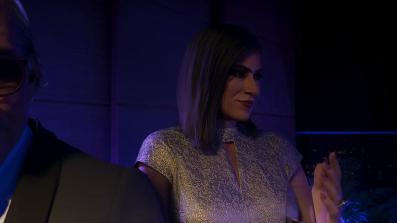 A smiling woman with a sleek bob in a shimmering silver dress stands in purple-blue light, with a man in sunglasses partly visible at the left edge.