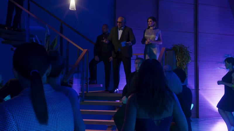 An older man in a dinner suit and sunglasses gestures from a staircase beside a woman in a silver dress, as a crowd watches in a blue-lit hall.