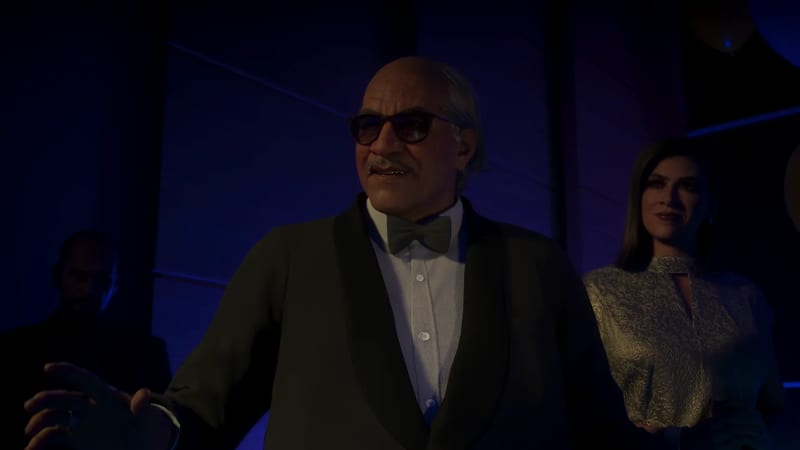 An older bald man with a moustache, tinted glasses, dinner jacket and bow tie gestures in a dim blue room, with a woman in a sparkly top behind him.