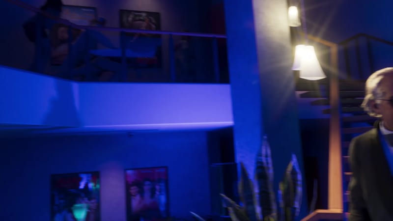 An older man in a dark jacket walks past a plant and a staircase inside a blue-lit, multi-level venue with posters on the walls.