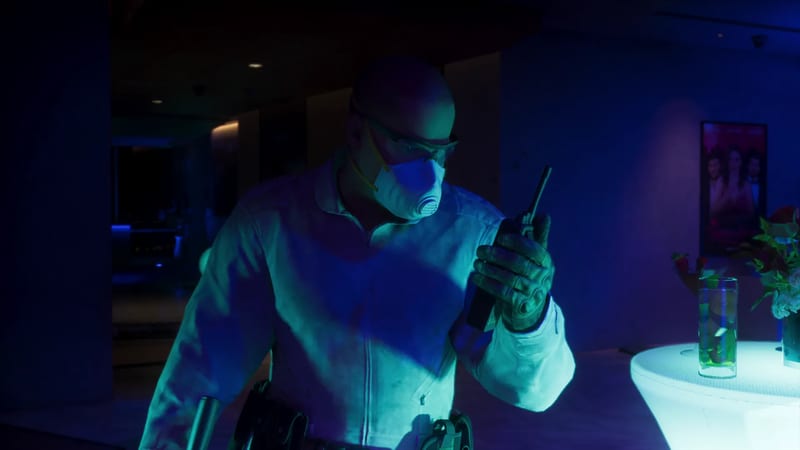 A man in a white jacket, face mask and safety glasses holds a walkie-talkie to his face in a dark room lit blue, beside a glowing white table.