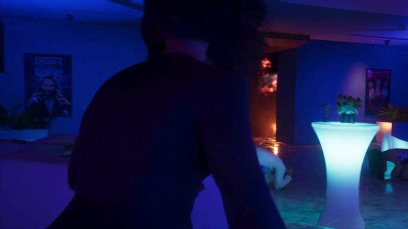 A dark-haired person seen from behind in a dim, blue-lit club or lounge, with a glowing white table and people crouching on the floor.