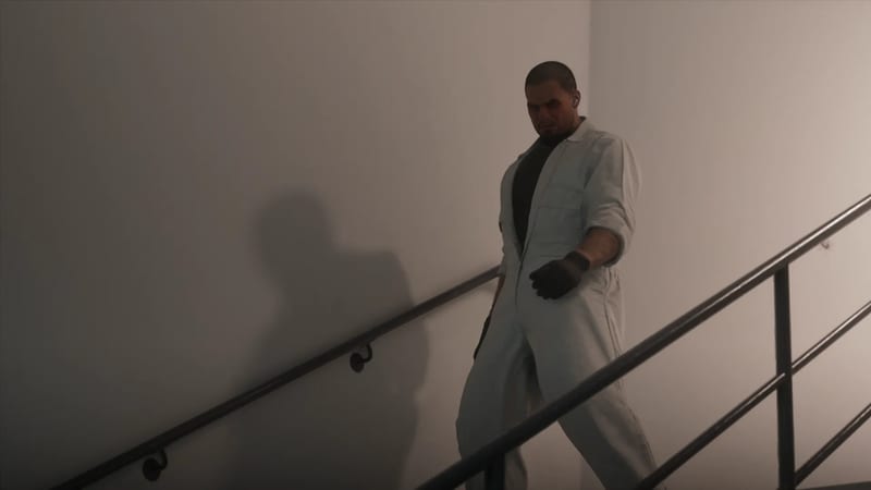 A man in a white boiler suit and dark gloves walks down a bare stairwell beside a metal handrail.