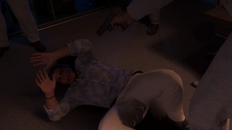 A man in a patterned shirt and light trousers lies on a carpeted floor with his hands raised while a person standing over him points a handgun.