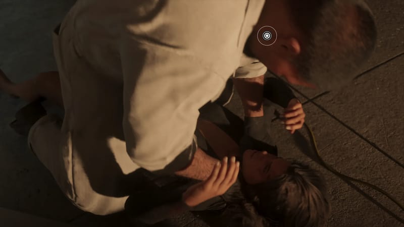 A bearded man in a cream shirt leans over a dark-haired woman lying on a paved floor, her hand raised, with a circular marker above them.