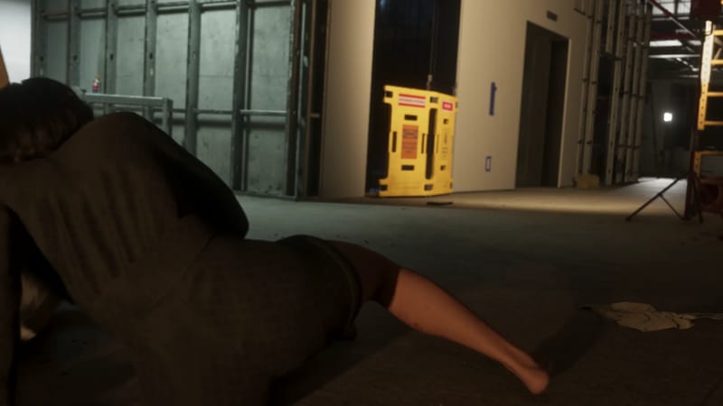 A dark-haired person in a dark jacket lies on a concrete floor in an unfinished building, with a yellow warning barrier behind them.