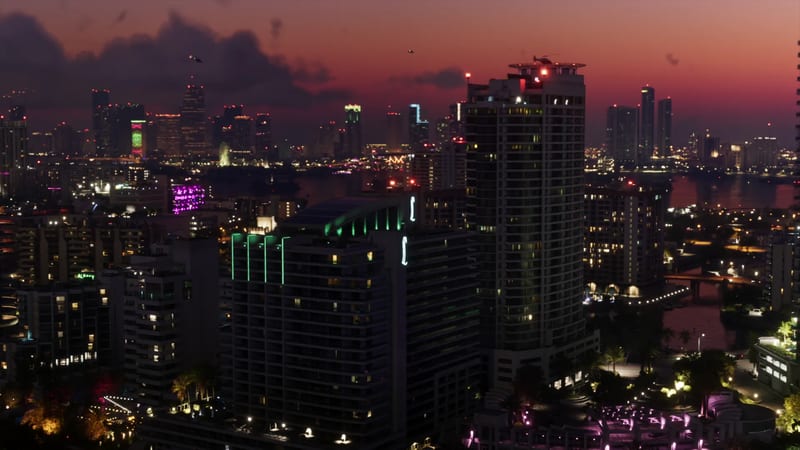 A dusk skyline of Vice City with lit high-rise towers, a helicopter above a rooftop helipad and a river beneath a red and purple sky.