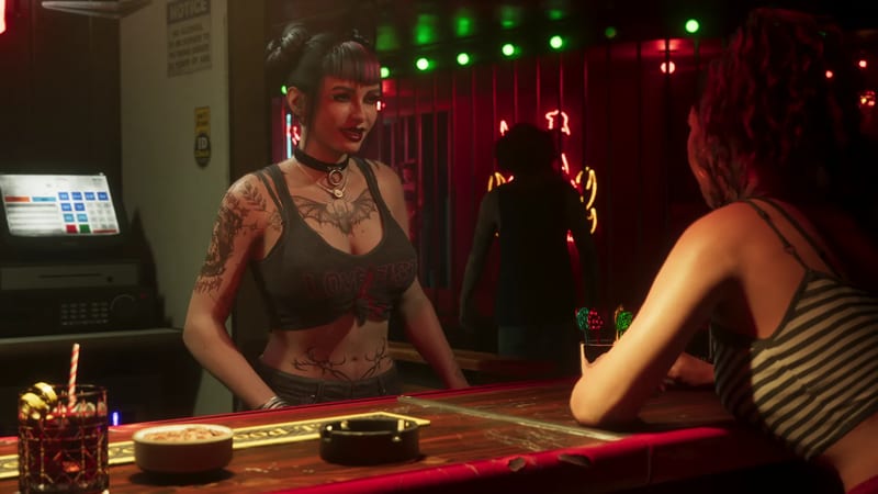 A tattooed woman with dark hair in buns, wearing a choker and grey top, talks to a woman in a striped top across a bar counter in a dimly lit, neon-lit bar.