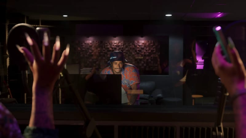 A smiling man in a bucket hat and orange patterned shirt looks at a screen in a dim recording studio, seen through glass past two raised, long-nailed hands.