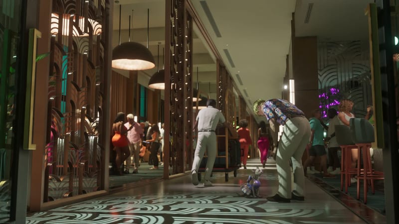 A man in a floral shirt bends to look at a small dog in a purple outfit as a porter pushes a luggage trolley down a busy art deco hotel corridor.