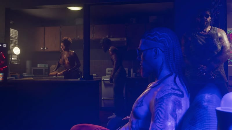 A shirtless tattooed man with long braids sits in blue light as two people work in a dim kitchen behind him and a bearded man stands nearby.