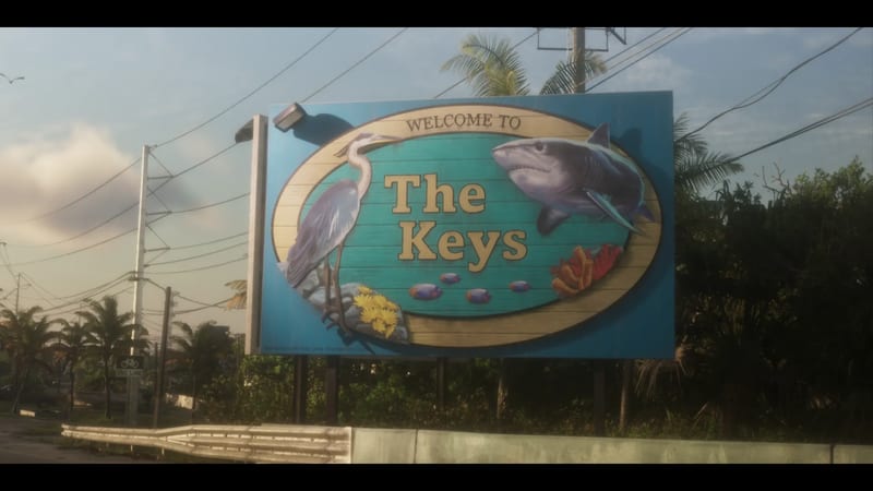 A large roadside billboard reading "Welcome to The Keys", showing a heron, a shark, fish and coral, with palm trees and power lines behind.