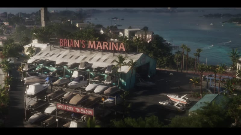 Boats on tall storage racks beside a teal boatyard shed with a red Brian's Marina sign, overlooking a turquoise bay and palm trees.