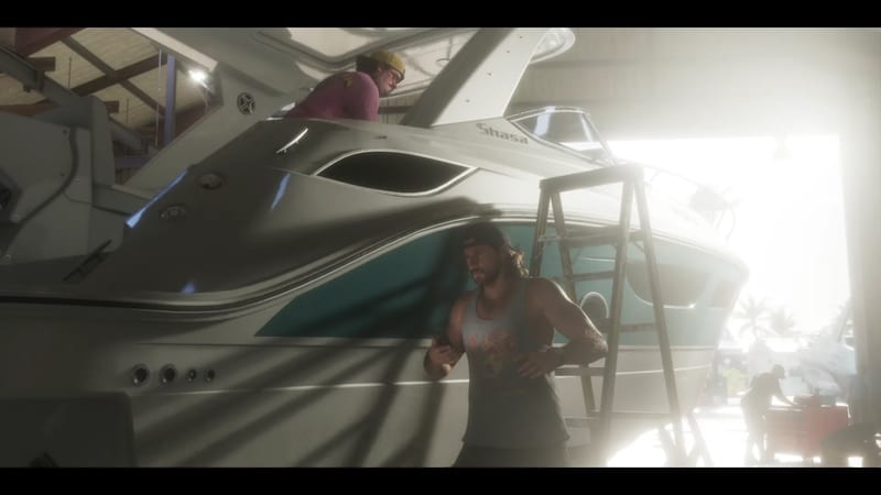 A bearded man in a grey vest leans on a white and teal motor yacht in a boatyard shed while another man in a yellow cap works on deck above.