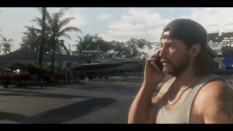 A bearded man in a backwards cap and grey vest talks on a phone in a sunny boatyard with boats and palm trees behind him.