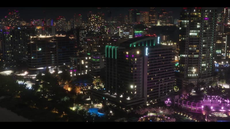 Aerial view of Vice City at night, with a lit-up hotel tower, neon-lit buildings and palm trees in the foreground.