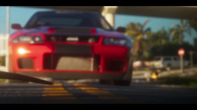 A red sports car with a black bonnet faces the camera on a road with yellow markings, beneath an overpass, with palm trees behind in bright sunshine.