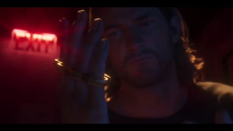 A bearded man with long hair holds up a gold knuckleduster in front of his face, with a glowing red exit sign blurred behind him.