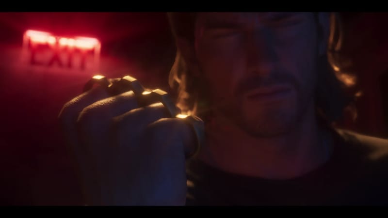 A long-haired, stubbled man in a dark top raises a fist wearing gold knuckledusters, with a glowing red EXIT sign blurred behind him in a dim room.