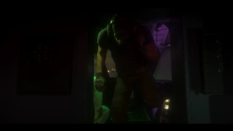 A man in a dark T-shirt and headset ducks through a dim helicopter doorway, lit green and purple, with city lights far below at night.