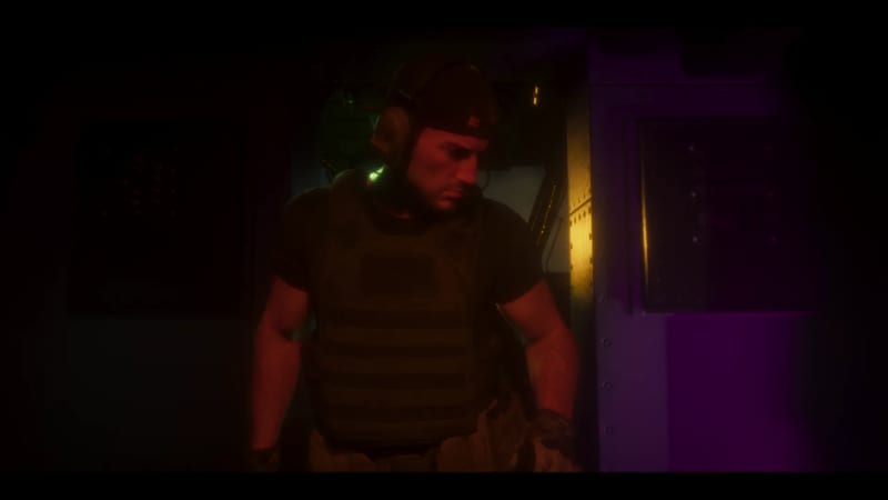 A bearded man in a backwards cap, headset and body armour stands in a dark metal compartment lit by red, purple and amber light.