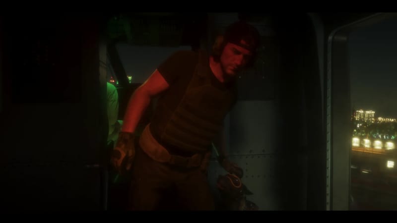 A bearded man in a backwards cap and tactical vest stands in the open door of a helicopter at night, a dog beside him and city lights outside.