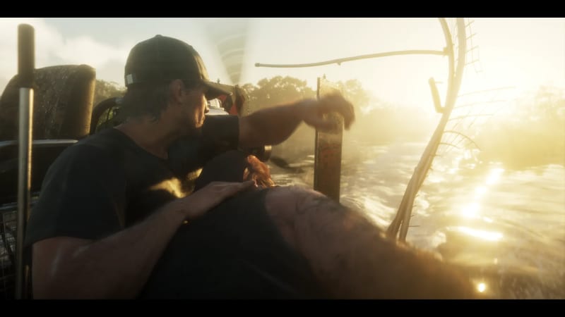 A man in a black cap and T-shirt reaches out from an airboat as it speeds across golden, sunlit swamp water.