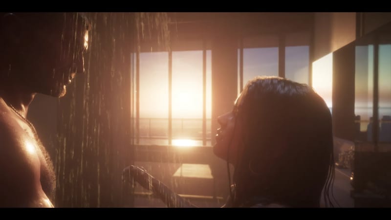A shirtless bearded man and a woman with long wet dark hair stand close together under running water, with a sunset glowing through large windows behind them.