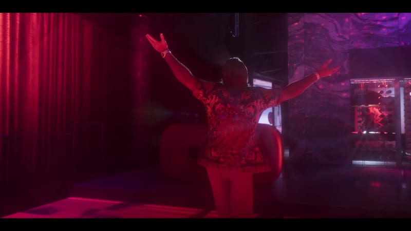 A man seen from behind raises both arms wide in a dark club lit in red and purple, wearing an ornate patterned shirt.