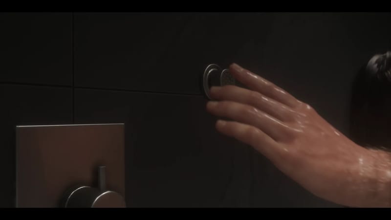 A wet hand reaches towards a small round button on a dark tiled shower wall, with a chrome tap handle at the bottom left.