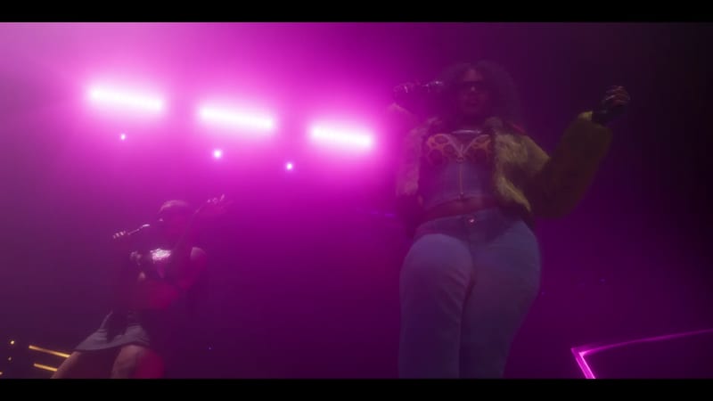 Two women perform on a stage lit in pink, one holding a microphone and wearing a fur jacket and jeans, the other crouching with a microphone.
