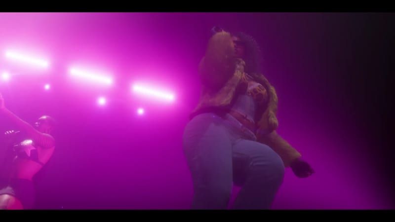 A woman with curly hair in a fur-trimmed jacket and light blue jeans dances on a stage lit with bright pink lights, with another performer at the left.