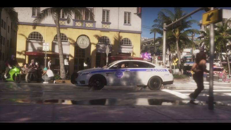 A white police car with a blue stripe drives along a wet sunlit street in Vice City, past palm trees, a yellow seafood restaurant and bikers.