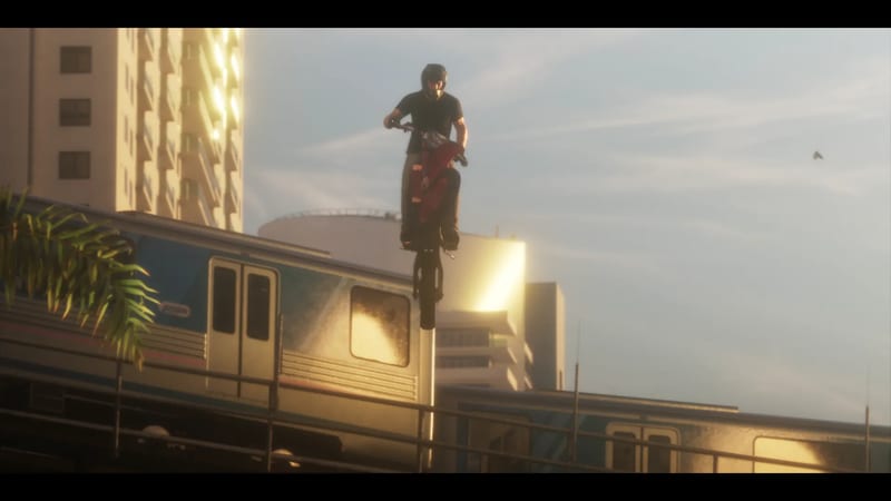 A helmeted man in a black T-shirt pops a wheelie on a red motorbike above an elevated train, with tall buildings behind in golden light.