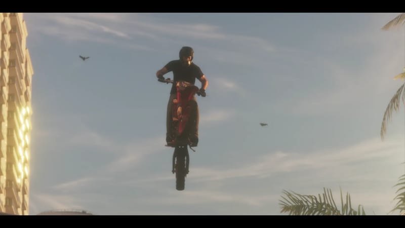 A rider in a black helmet and black T-shirt launches a red dirt bike high into the air against a hazy blue sky, with a tall building and palm fronds nearby.