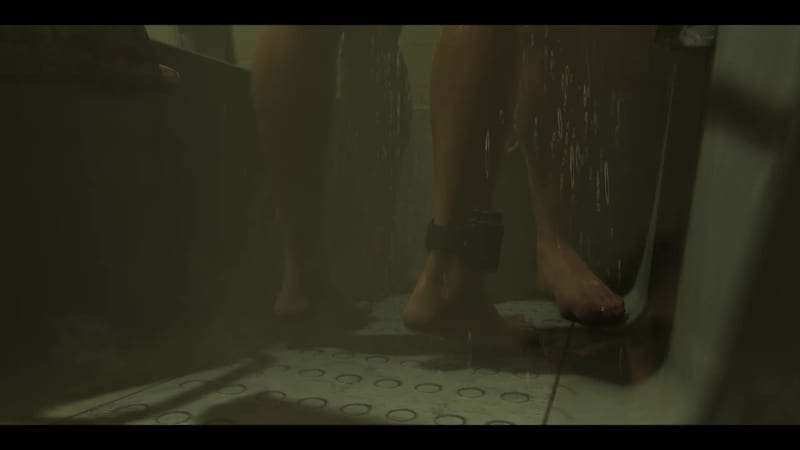 Two people's bare legs and feet stand in a wet, dimly lit shower, one with a black electronic tag fastened around the ankle.