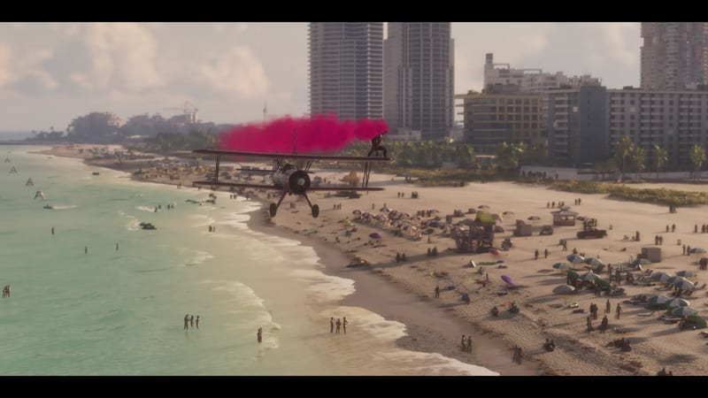 A red biplane trailing pink smoke flies low over Ocean Beach, a person standing on its upper wing, with sunbathers below and tall apartment blocks behind.