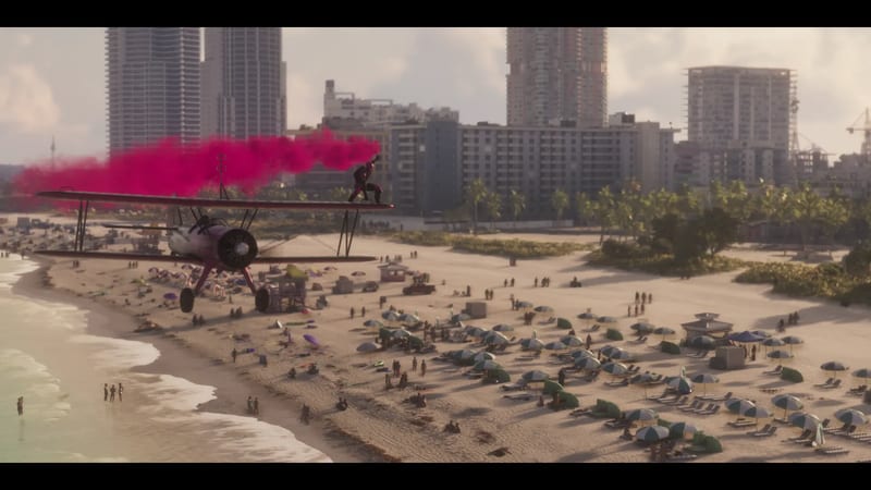 A person stands on the wing of a biplane trailing pink smoke over a busy Ocean Beach, with high-rise buildings behind.