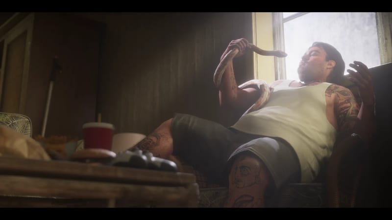 A heavily tattooed man in a white vest and shorts reclines in an armchair by a bright window, holding a pale snake that is draped across his chest.