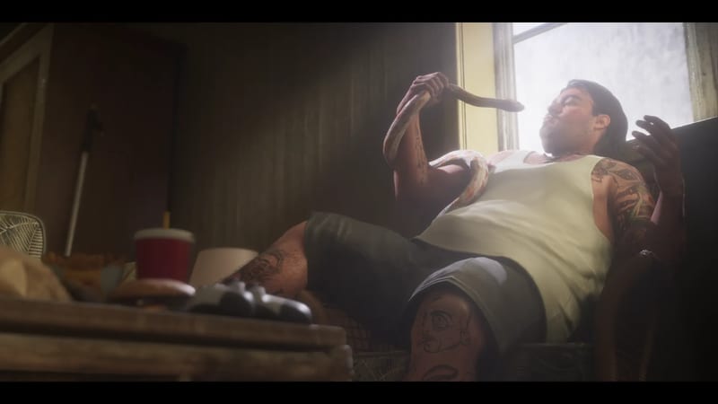 A heavily tattooed man in a white vest and grey shorts reclines in a chair by a bright window, holding a pale snake draped over his shoulder.