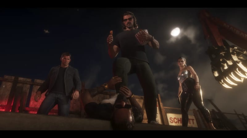 A bearded man in sunglasses eats fried chicken and rests his boot on a man lying on the ground, as two onlookers watch at night on a building site.