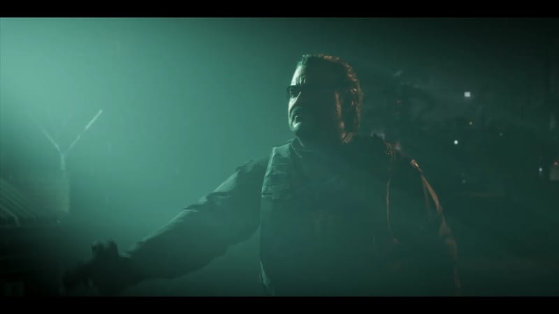 A heavyset man in glasses and a dark tactical vest stands with one arm stretched out in green-lit mist at night, beside a tall fence.