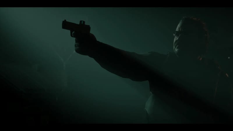 A man in glasses, mostly in shadow, aims a handgun with a small sight on top out of frame to the left in a dark, green-tinted space.