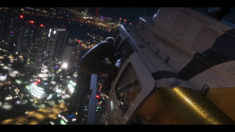A man in dark clothes clings to the outside of a white and yellow helicopter high above a brightly lit city at night.
