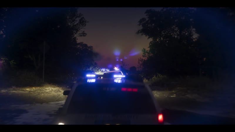 A white police pickup with flashing blue lights leads other police vehicles along a wet, muddy road through dark trees at night.