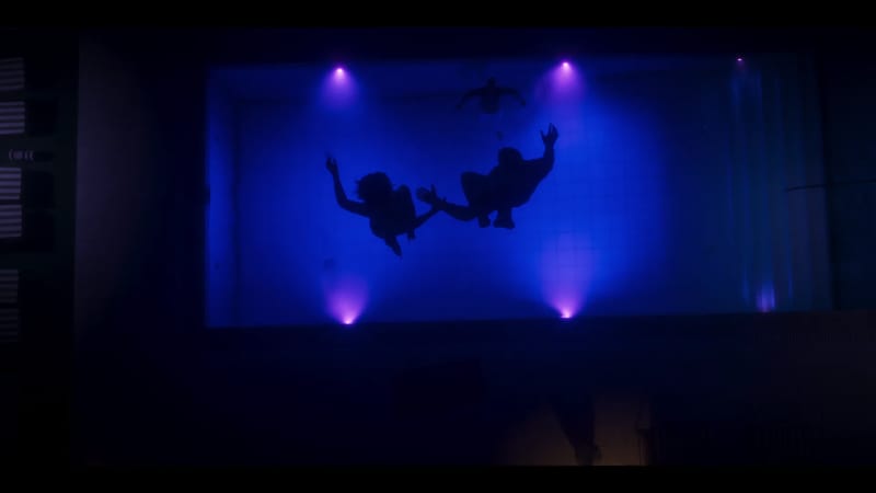 Silhouettes of two people float underwater in a tiled pool lit deep blue, with a third swimmer near the surface and purple lights around the edges.