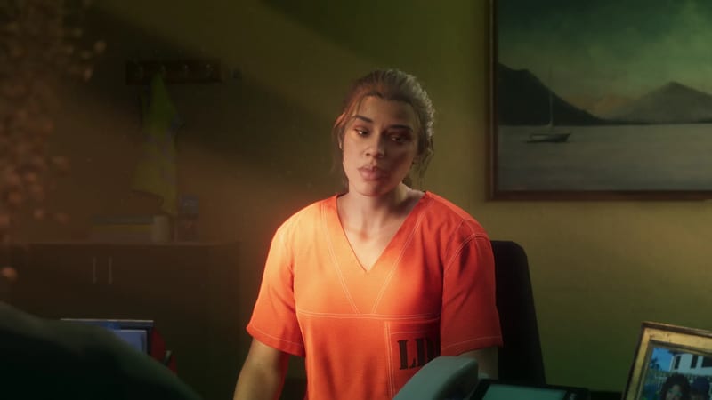 Lucia Caminos, in an orange prison top, sits at a desk in a dim office with a framed lake painting behind her.